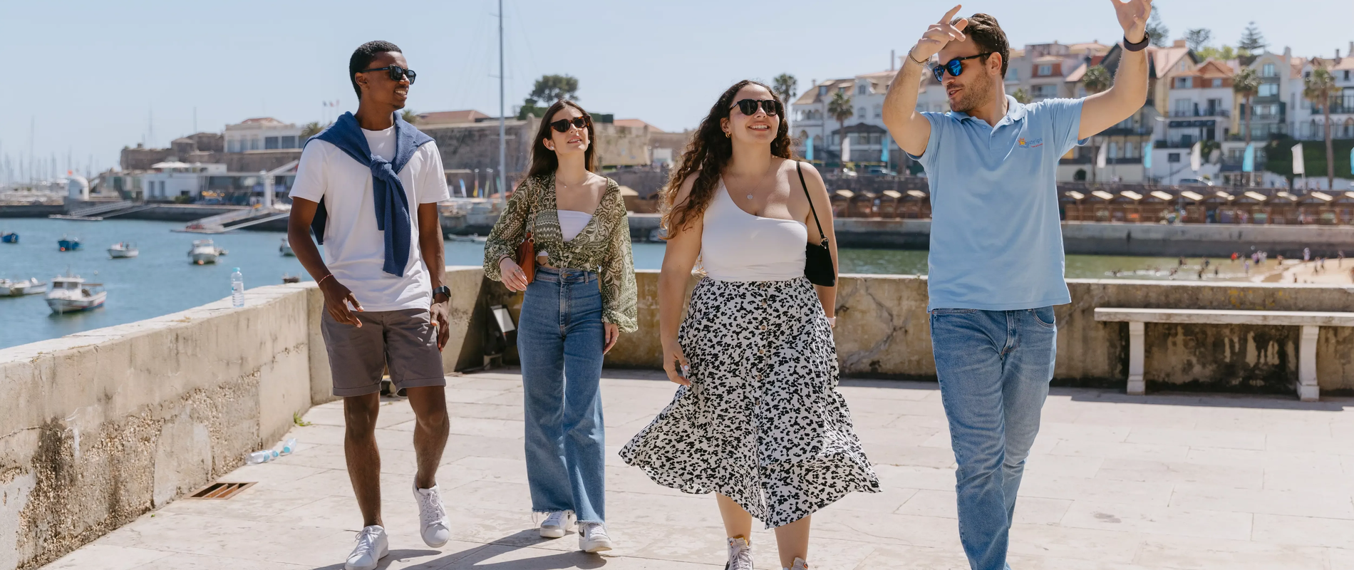 Group of people enjoying a guided walking tour along the sunny coast of Cascais, Portugal, with boats and colorful houses in the background