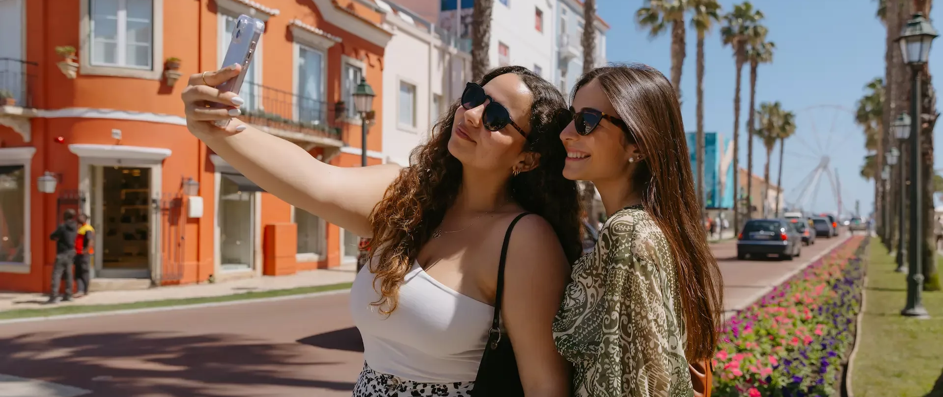 Two young women taking a selfie in the heart of Cascais, Portugal, on a bright sunny day. They are smiling and wearing sunglasses, standing near colorful flower beds and palm trees, with a vibrant street scene and Ferris wheel visible in the background. A picturesque moment in this coastal town known for its beauty and charm.