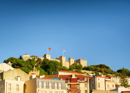 View of São Jorge Castle in Lisbon, Portugal, with flags waving above historic walls and colorful houses below.