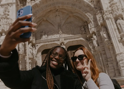 Two tourists taking a selfie in front of the ornate entrance of Jerónimos Monastery in Lisbon, Portugal.