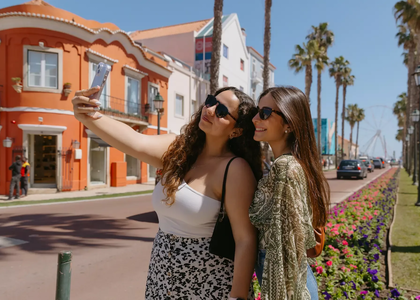 Two young women taking a selfie in the heart of Cascais, Portugal, on a bright sunny day. They are smiling and wearing sunglasses, standing near colorful flower beds and palm trees, with a vibrant street scene and Ferris wheel visible in the background. A picturesque moment in this coastal town known for its beauty and charm.