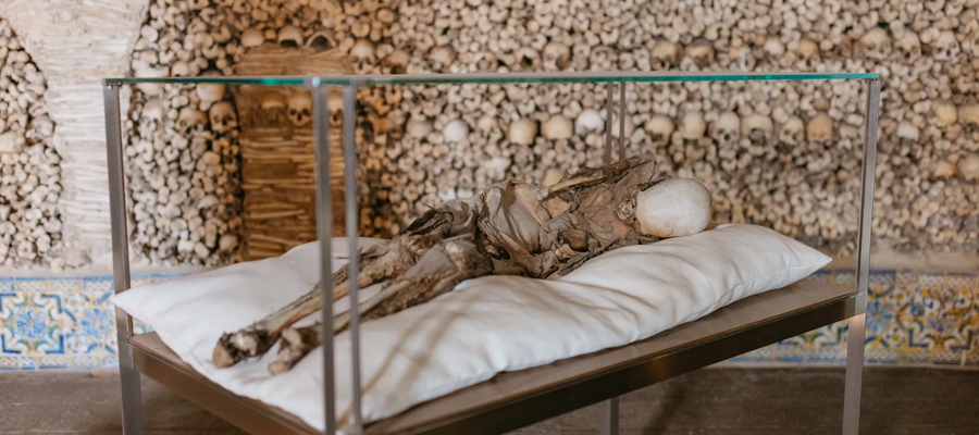 Interior view of the Chapel of Bones in Alcácer do Sal, Portugal, featuring a mummified body on display and walls covered with human skulls and bones.