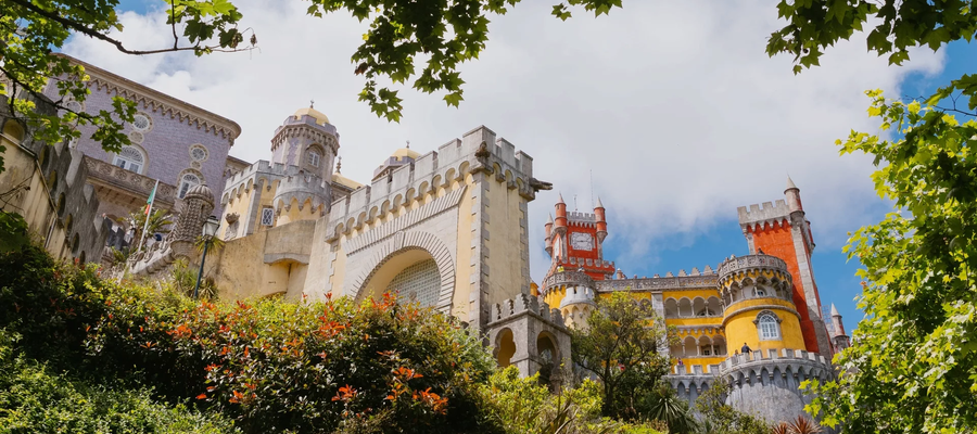 Colorful view of the Pena Palace in Sintra, Portugal, surrounded by lush greenery under a partly cloudy sky.