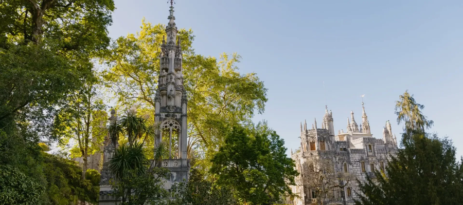 View of the Gothic tower and main palace of Quinta da Regaleira in Sintra, Portugal, surrounded by lush greenery under a clear blue sky.