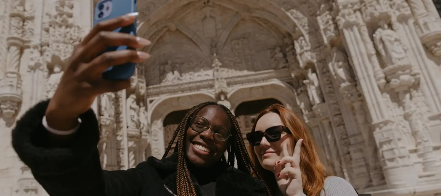 Two tourists taking a selfie in front of the ornate entrance of Jerónimos Monastery in Lisbon, Portugal.