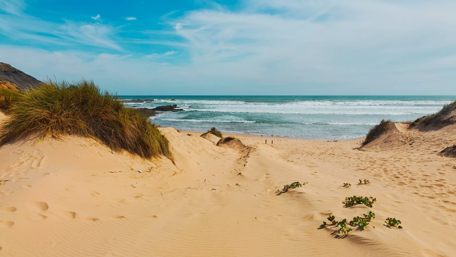 “Praia da Amoreira in Aljezur, Portugal sandy dunes, grassy vegetation and the Atlantic Ocean under a partly cloudy sky.”