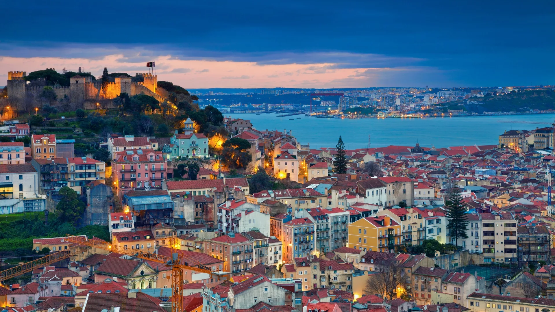 Scenic view of Lisbon at sunset, with São Jorge Castle illuminated above the historic Alfama district and the Tagus River in the background.