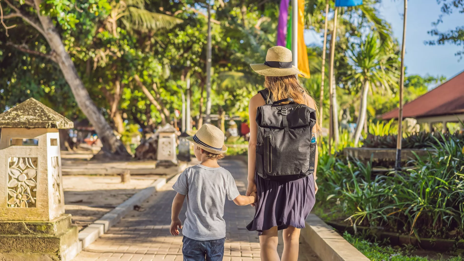 Mother and young son walking hand in hand down a sunny tropical path, both wearing straw hats, with a backpack and lush greenery around them.