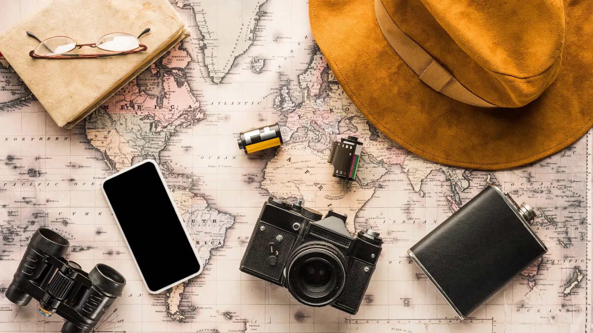 Flat lay of vintage travel essentials on an old world map, including a retro camera, smartphone, film rolls, notebook with glasses, flask, binoculars, and a wide-brimmed suede hat.