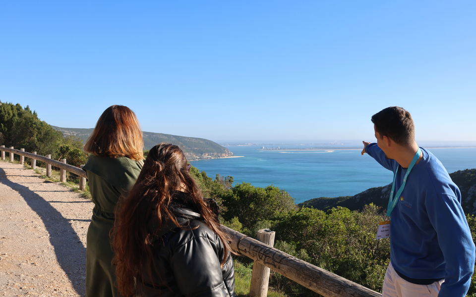 Tour guide pointing towards the Sado Estuary and Tróia Peninsula from a scenic viewpoint in the Arrábida Mountains, with two visitors observing.
