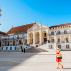 Female tourist in a red dress walking across the courtyard of the University of Coimbra, Portugal, under a bright blue sky.