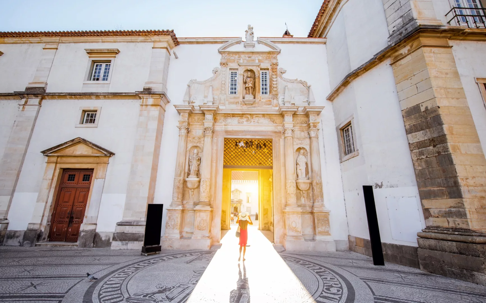 Main gate of the University of Coimbra (Porta Férrea) lit by the sun, with stone statues and mosaic pavement.