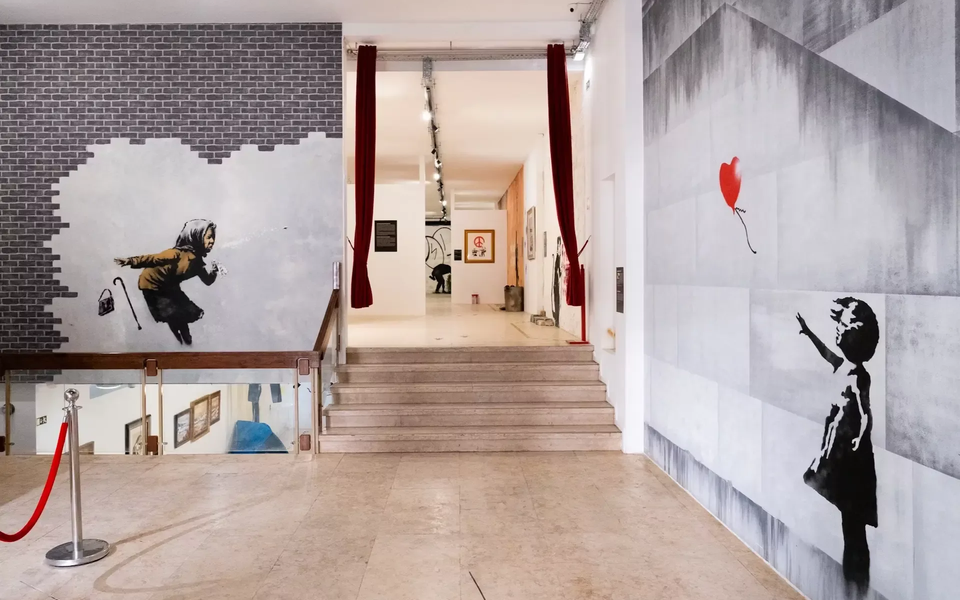 Interior view of The World of Banksy exhibition in Lisbon featuring two iconic murals — “Girl with Balloon” and “Achoo!!” — displayed on adjacent walls near the main staircase.