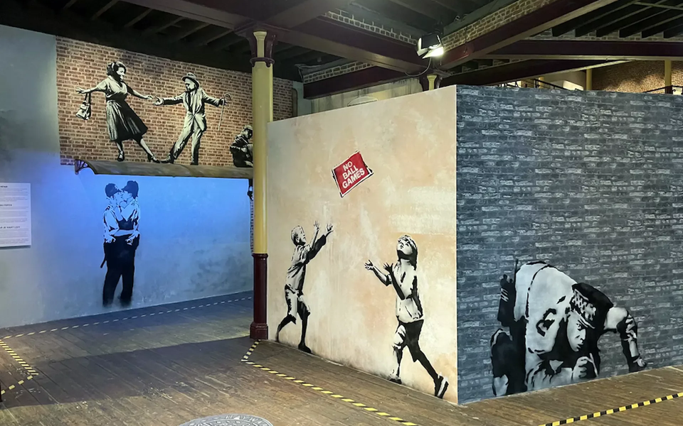 Gallery view of several Banksy murals, including children playing under a “No Ball Games” sign, two policemen kissing, and a dancing couple on a brick wall, all presented in a recreated urban environment.