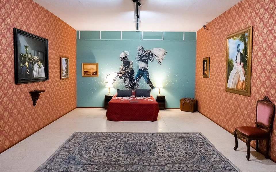 Recreation of a bedroom from Banksy’s Walled Off Hotel, featuring a stenciled mural of two figures in a pillow fight, surrounded by vintage-style furniture, classic paintings, and a richly patterned carpet.