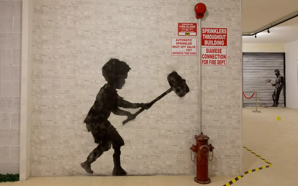 Indoor wall art installation from the World of Banksy in Lisbon featuring a stenciled silhouette of a child with a sledgehammer near a real fire hydrant, blending street art with real-world elements. Another Banksy-style piece is visible in the background.