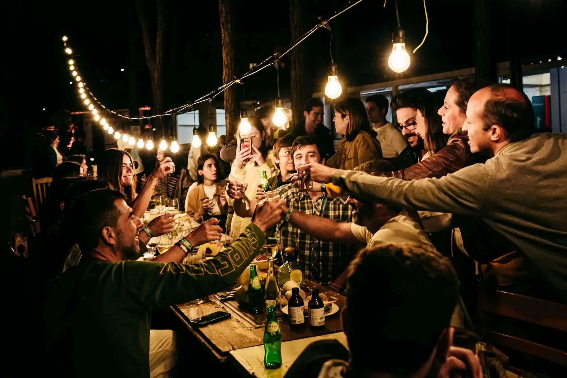 People gathered around a long wooden table at night, sharing drinks and toasting under warm hanging light bulbs at an outdoor food festival.
