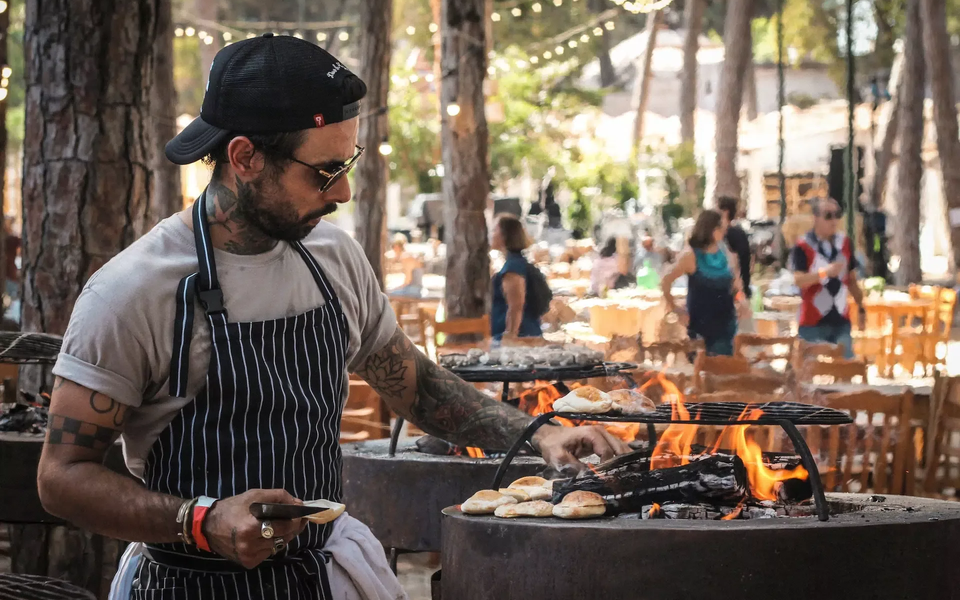 Tattooed chef grilling bread over an open flame at an outdoor food festival set in a pine forest with guests in the background.