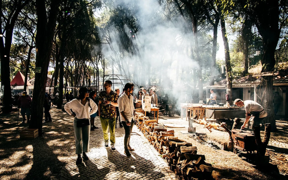People walking through a sunlit forest food festival with smoke rising from open-fire cooking areas and firewood stacked along a path.