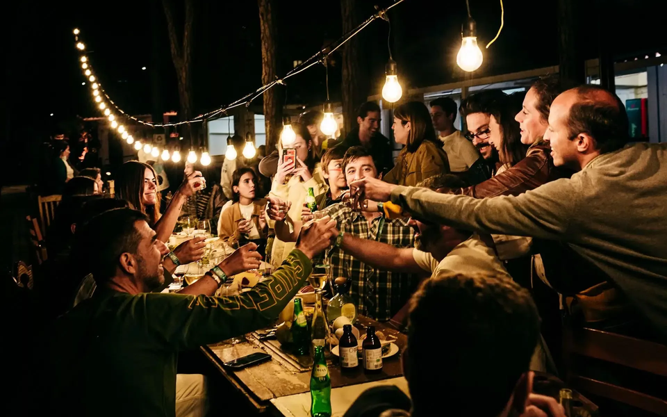 People gathered around a long wooden table at night, sharing drinks and toasting under warm hanging light bulbs at an outdoor food festival.