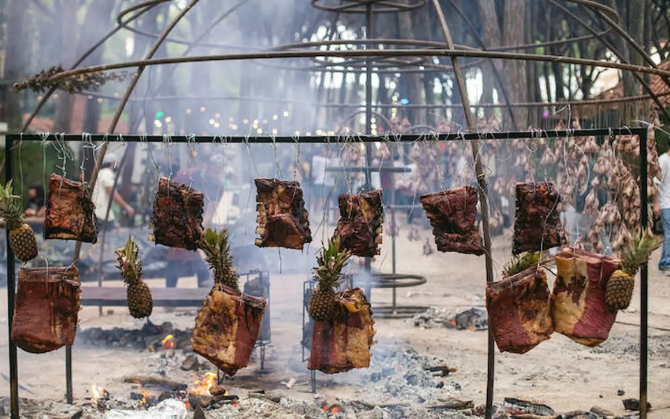 Outdoor fire-cooking scene with large cuts of meat and whole pineapples hanging above an open flame at a rustic food festival or barbecue event, with people and forest surroundings in the background.