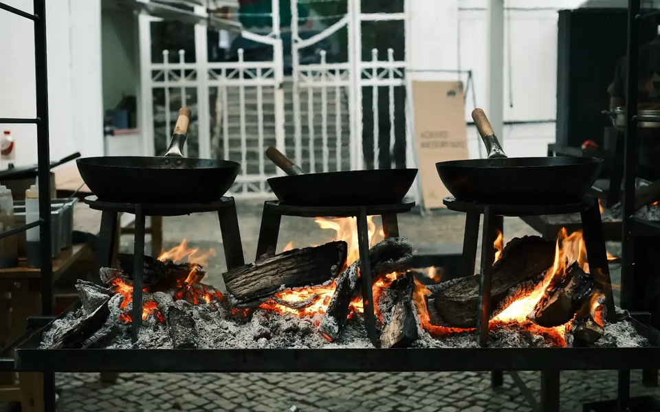 Close-up of three cast iron pans on metal stands, cooking over an open wood fire with glowing embers and flames, set in an outdoor urban environment with cobblestone ground.