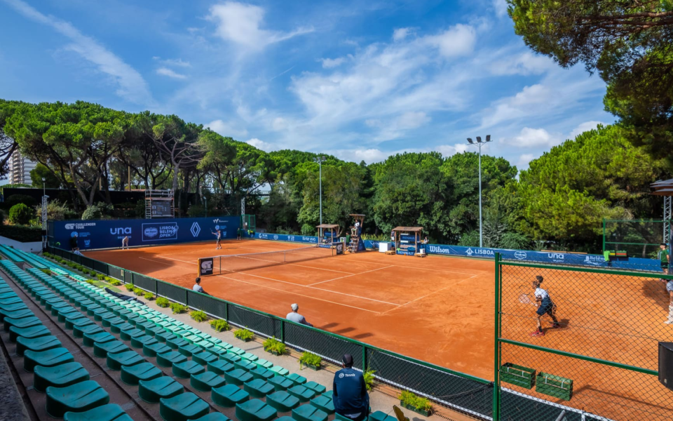 Lisboa Belém Open tennis court at CIF venue in Lisbon, with a red clay surface, surrounded by lush pine trees. The image shows a professional tennis match in progress, with a crowd seated on bleachers and a clubhouse in the background. Ideal for sports tourism, ATP/WTA events, and cultural sports festivals in Portugal.