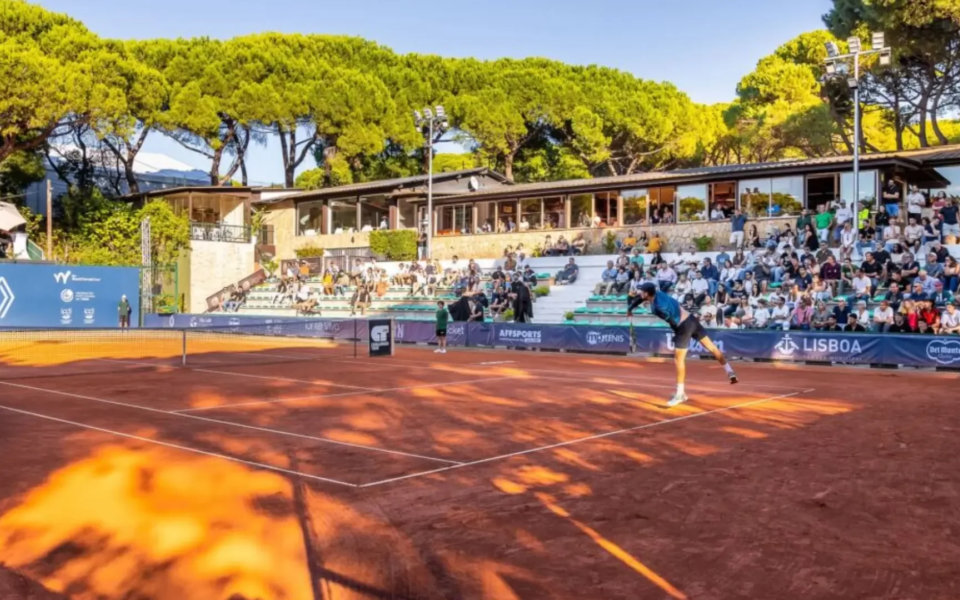 The tennis court at CIF in Lisbon, Portugal, during the Lisboa Belém Open tournament. The scene shows a professional match in progress on a red clay court, with spectators seated in shaded bleachers and pine trees in the background. A modern clubhouse overlooks the action, capturing the blend of sport, nature, and vibrant local atmosphere.