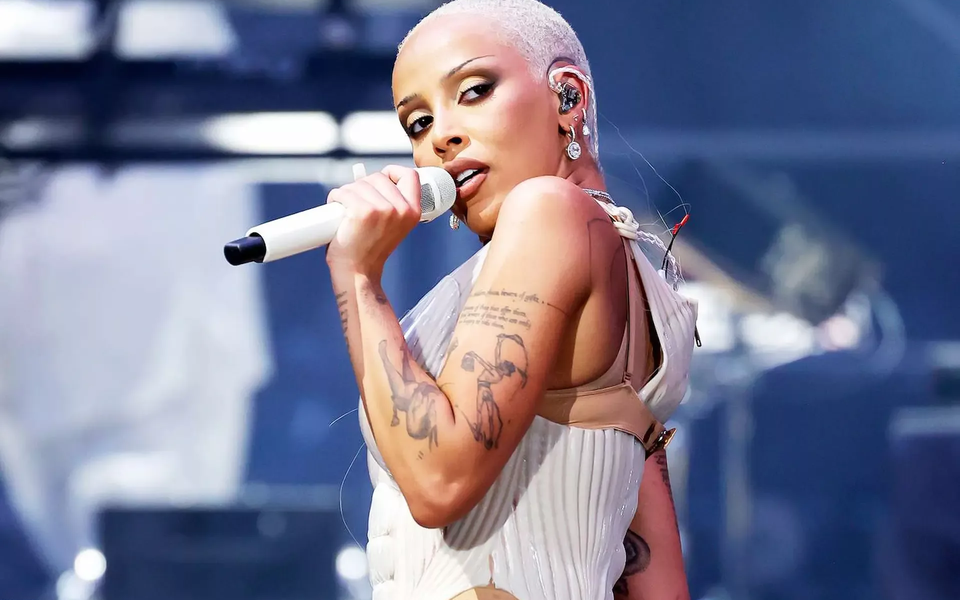 Doja Cat singing into a white microphone during a live concert, in a close-up shot showing her tattoos and stage costume under bright lights