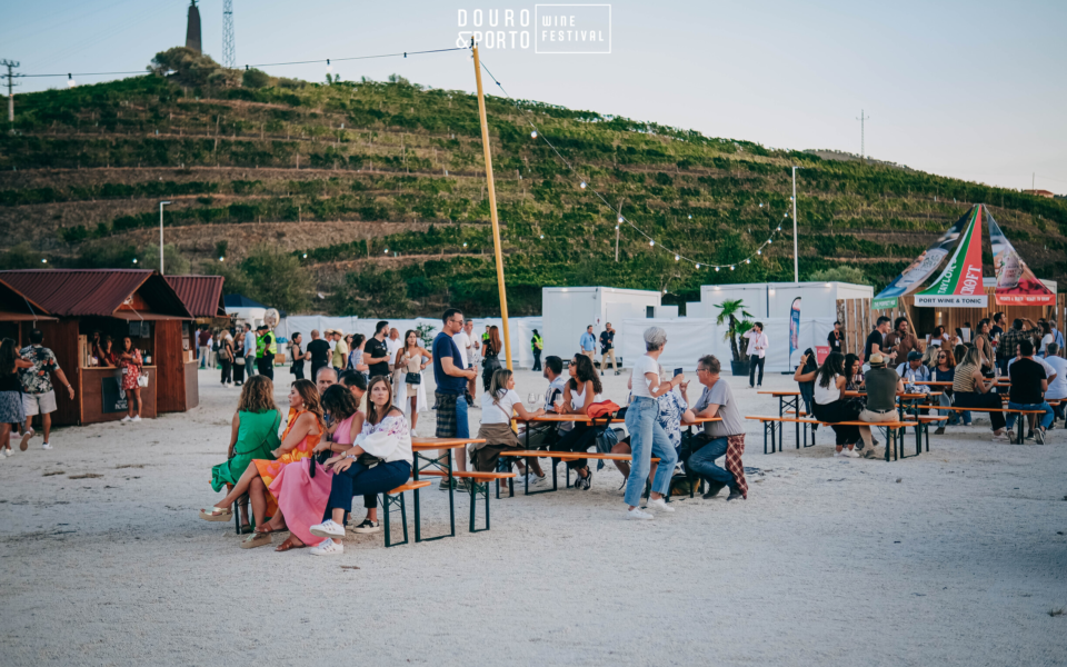 Daytime scene of Douro & Porto Wine Festival with people, booths, and vineyard hills in the background