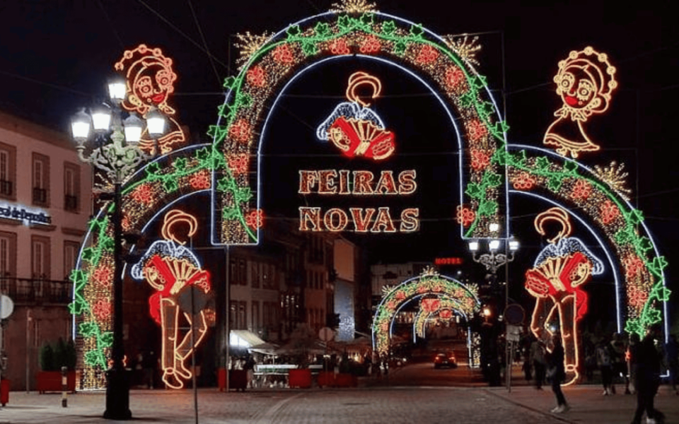 Night view of the illuminated arches at Feiras Novas festival in Ponte de Lima, Portugal. Decorative lights depict traditional figures with accordions and flowers, creating a festive atmosphere in the historic town center.