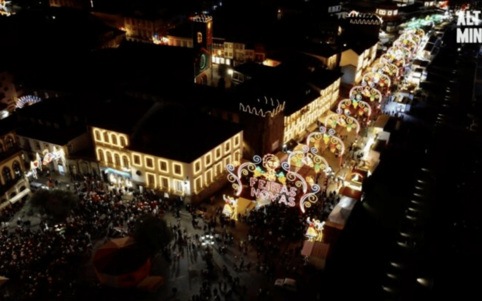 Aerial night view of the Feiras Novas festival in Ponte de Lima, Portugal, showcasing illuminated arches and a large crowd in the historic town center. Traditional decorative lights and festive atmosphere dominate the scene.