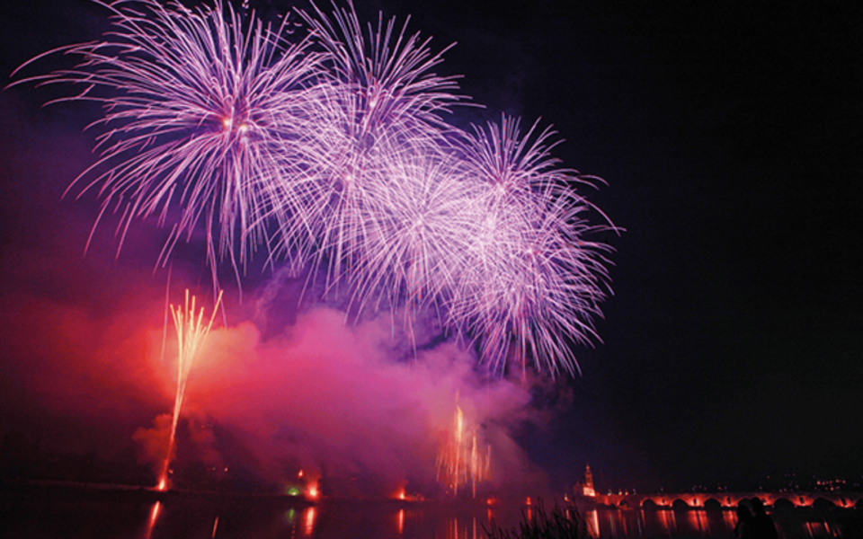 Vibrant fireworks display lighting up the night sky over Ponte de Lima during the Feiras Novas festival. The colorful bursts are reflected in the river, with the illuminated historic bridge and church tower in the background, and silhouettes of spectators in the foreground.