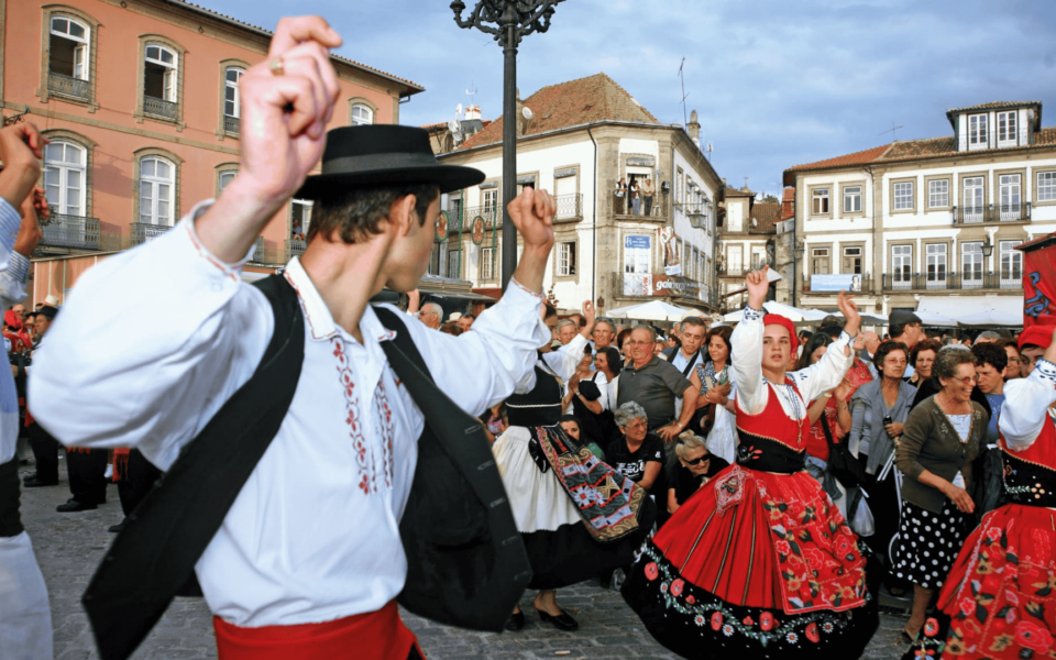 Traditional folk dancers in vibrant regional costumes performing in the main square of Ponte de Lima during the Feiras Novas festival. The image captures the energy of the crowd, embroidered details of the red skirts, and the historical architecture in the background.