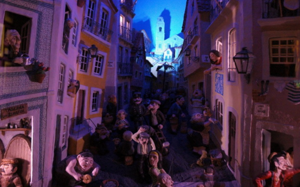 Artistic installation representing Lisbon's Popular Festivities, featuring clay miniatures of traditional neighborhood life, with colorful buildings, decorated balconies, and lively street scenes under a blue night sky.