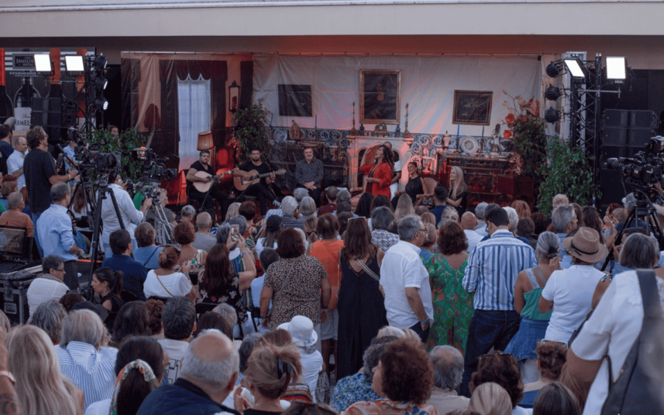 Outdoor Fado concert in Lisbon, Portugal, featuring a live performance with traditional Portuguese guitarists and a Fado singer on a decorated stage. The audience is seated and standing, enjoying the show in a cozy, intimate setting with media coverage and professional lighting.