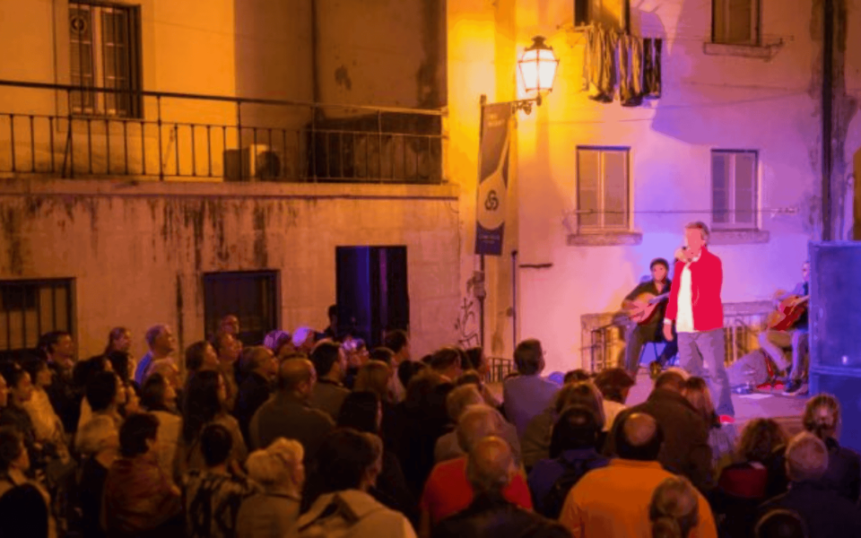 Evening Fado performance in the streets of Alfama, Lisbon. A male Fado singer is accompanied by two traditional Portuguese guitarists, performing on a small stage illuminated by warm yellow and purple lights. A large crowd watches the cultural street concert in an authentic Lisbon neighborhood.