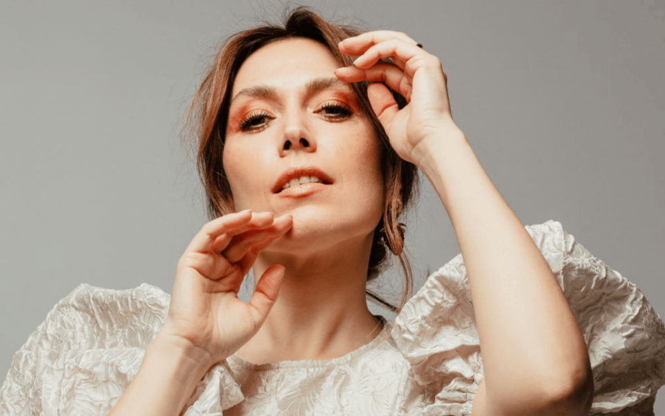 Close-up official portrait of Ana Bacalhau, Portuguese singer, in a cream textured blouse. She poses with raised hands in front of a neutral background, exuding elegance and confidence. Ideal for use in music festival promotions, artist profiles, and cultural event materials.