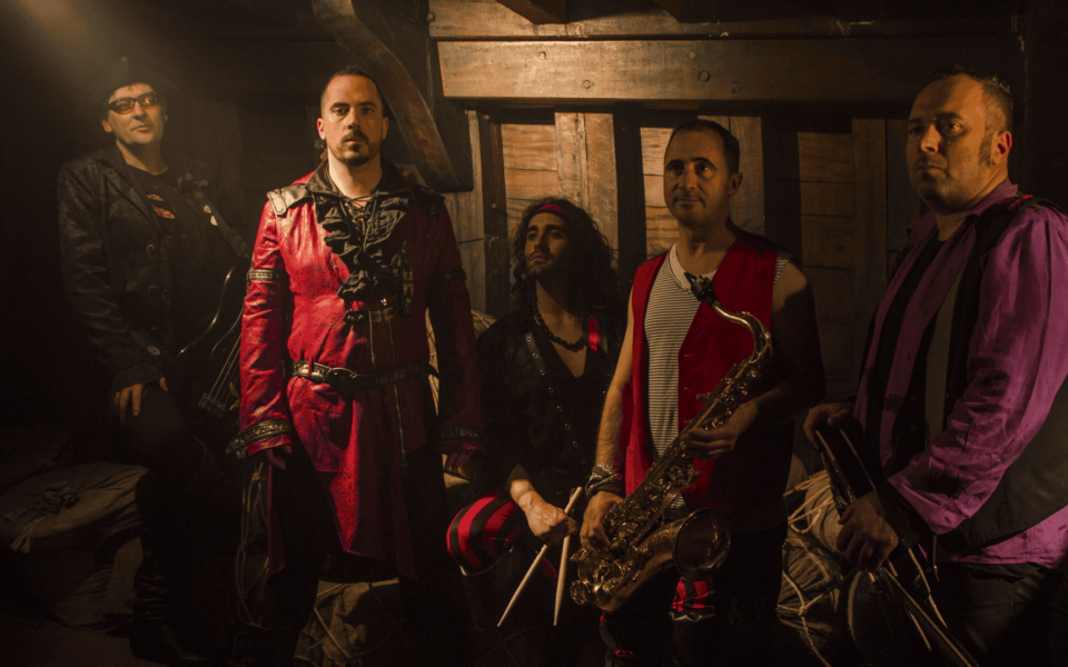 Rocktulo band members in dramatic pirate-themed costumes inside a rustic wooden ship-like setting, holding musical instruments including a saxophone, guitar, and drumsticks. Ideal image for promoting themed music festivals, theatrical rock performances, and alternative band lineups in Portugal.