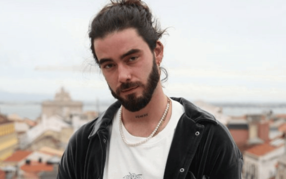 Close-up portrait of Portuguese singer SYRO, featuring a soft expression, wearing a black velvet shirt and pearl necklace with Lisbon rooftops in the background. Ideal for concert promotions, press kits, and music festival websites highlighting modern Portuguese music talent.