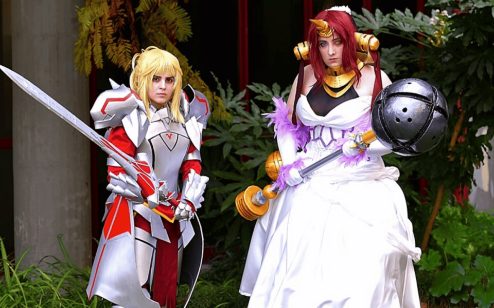 Two cosplayers dressed in elaborate fantasy armor and gowns pose at Iberanime 2025, Portugal’s leading Japanese pop culture event. One holds a sword and the other wields a large mace, surrounded by lush greenery near the event venue.
