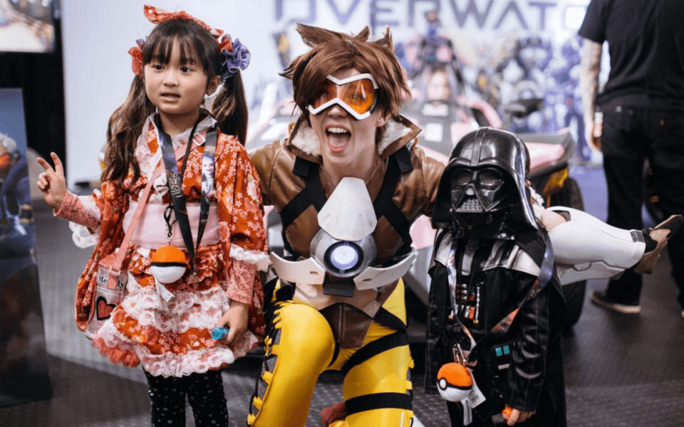 Children and an adult cosplaying at Iberanime 2025 in Portugal. One child wears a red kimono-style costume, another is dressed as Darth Vader, and the adult is cosplaying as Tracer from Overwatch. A fun and inclusive scene from a pop culture and anime convention.