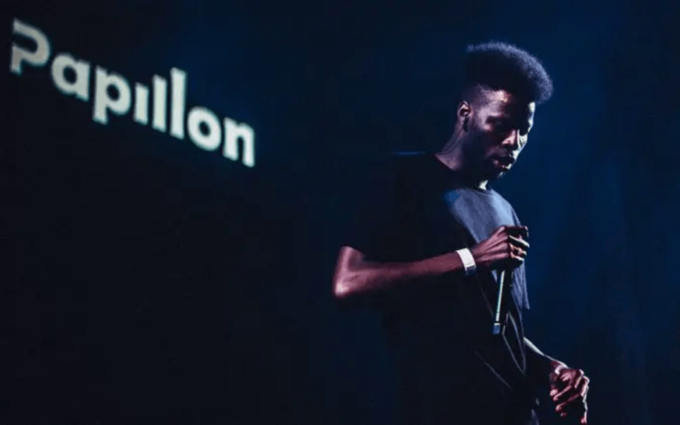 Portuguese rapper Papillon performing live on stage, holding a microphone under dramatic lighting, with his name projected in the background. Dark ambiance emphasizes the artist's presence during the concert.