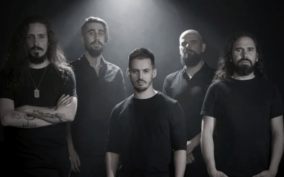 Promotional photo of a Portuguese rock or metal band with five male members, dressed in black, standing under moody lighting. Strong shadows and dark background highlight the group's intense and artistic image for an upcoming live concert.