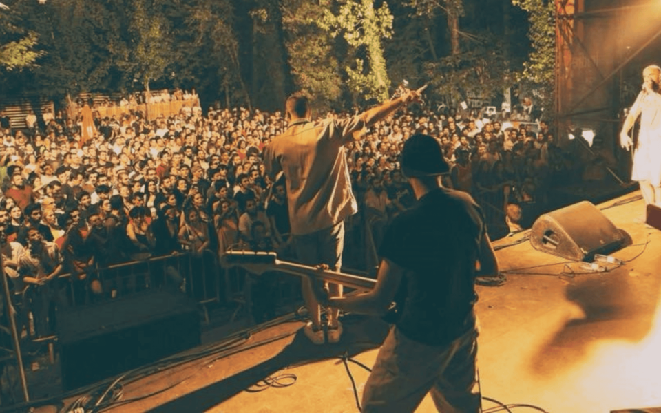 Live concert scene at Indie Music Fest 2025 in Baltar, Paredes. The image captures musicians performing on stage with a cheering crowd in a forested outdoor venue, under warm lighting.