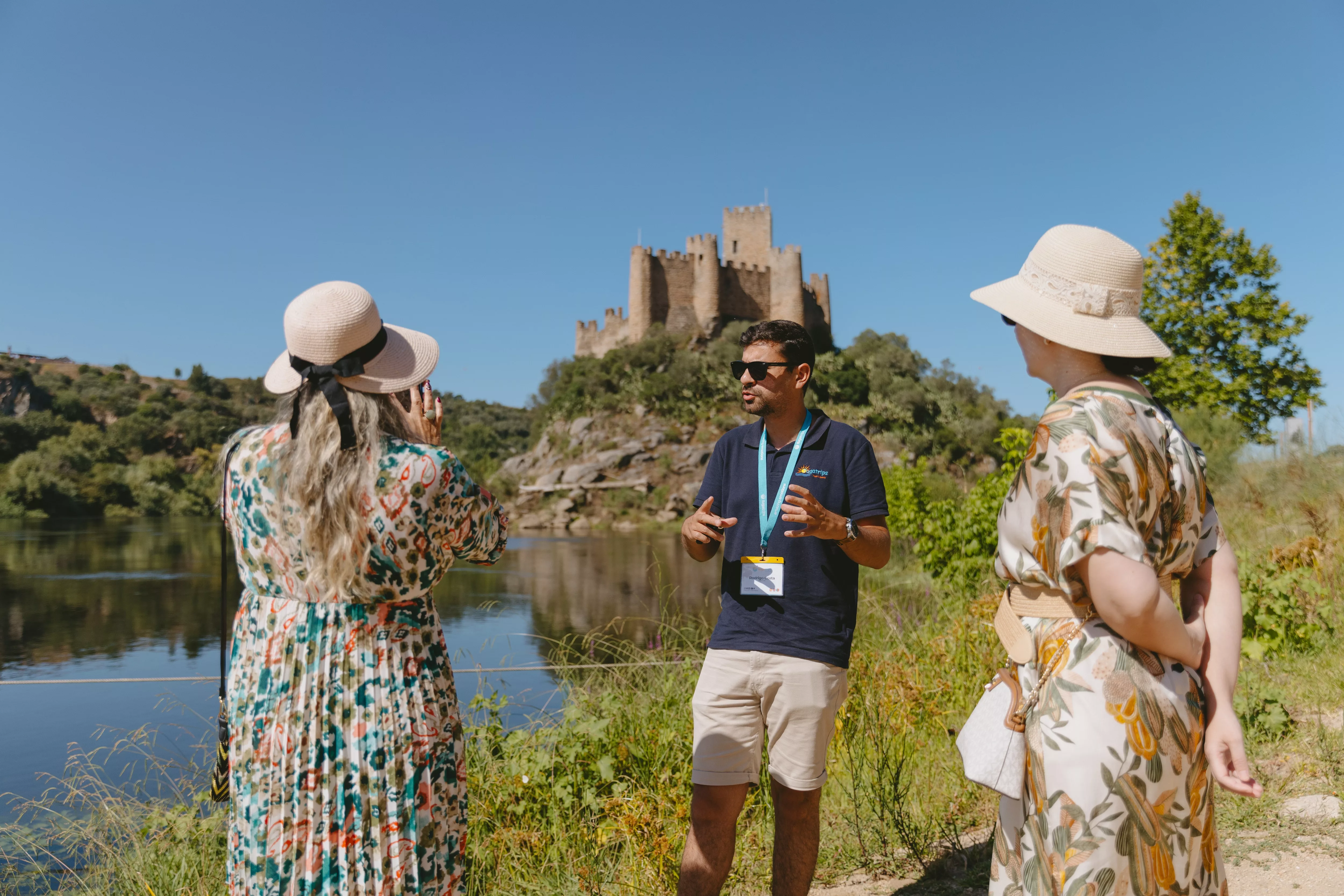 Tour guide explaining the history of Almourol Castle in Tomar, Portugal, with visitors by the river.
