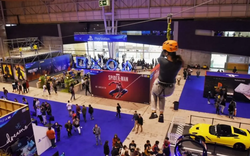 Indoor zip line attraction at Lisbon Games Week 2025, featuring a participant mid-air above a packed PlayStation exhibition zone. The scene includes promotional booths for Spider-Man and Call of Duty, a vibrant gaming crowd, and a yellow sports car display, highlighting the event’s mix of interactive experiences and blockbuster gaming titles.