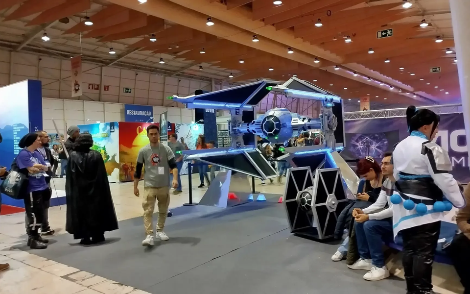 Star Wars-themed installation at Lisbon Games Week 2025, featuring a full-size replica of a TIE Fighter illuminated with LED lights. Visitors, including cosplayers and fans, gather around the exhibit in the indoor venue of FIL, enjoying one of the most iconic sci-fi attractions of the event.