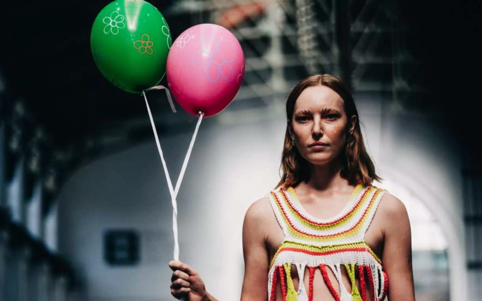 Female model on the runway wearing a colorful crochet dress and holding two balloons, during a contemporary fashion show