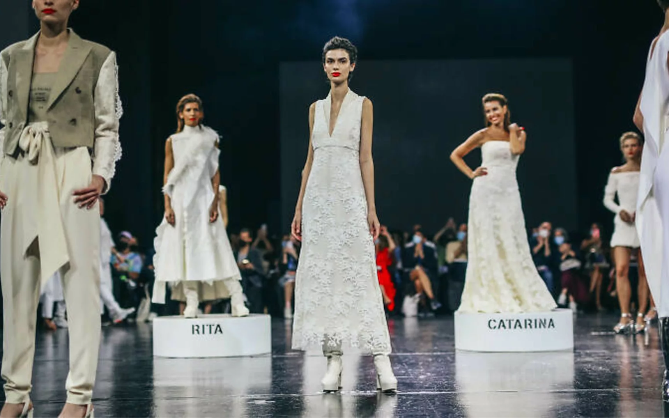 Models wearing elegant white dresses on stage with platforms labeled Rita and Catarina during a fashion show in Lisbon